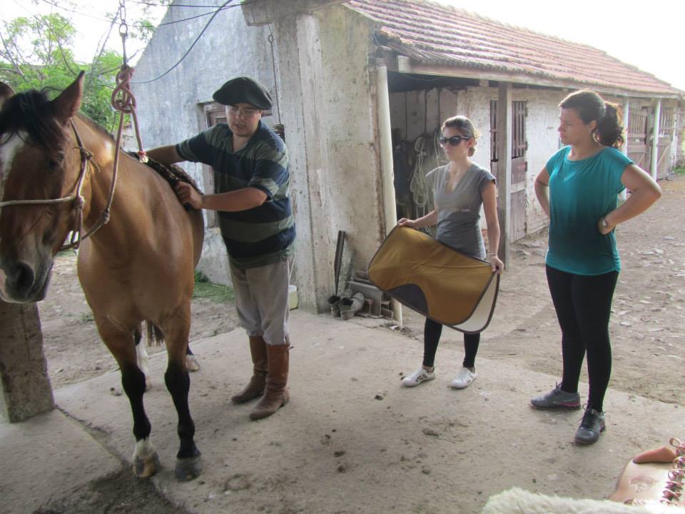 À esquerda, um cavalo está preso por rédeas, que um homem com boina segura; ao lado dele, uma moça se aproxima com um  tecido nas mãos, e outra moça observa, com as mãos na cintura