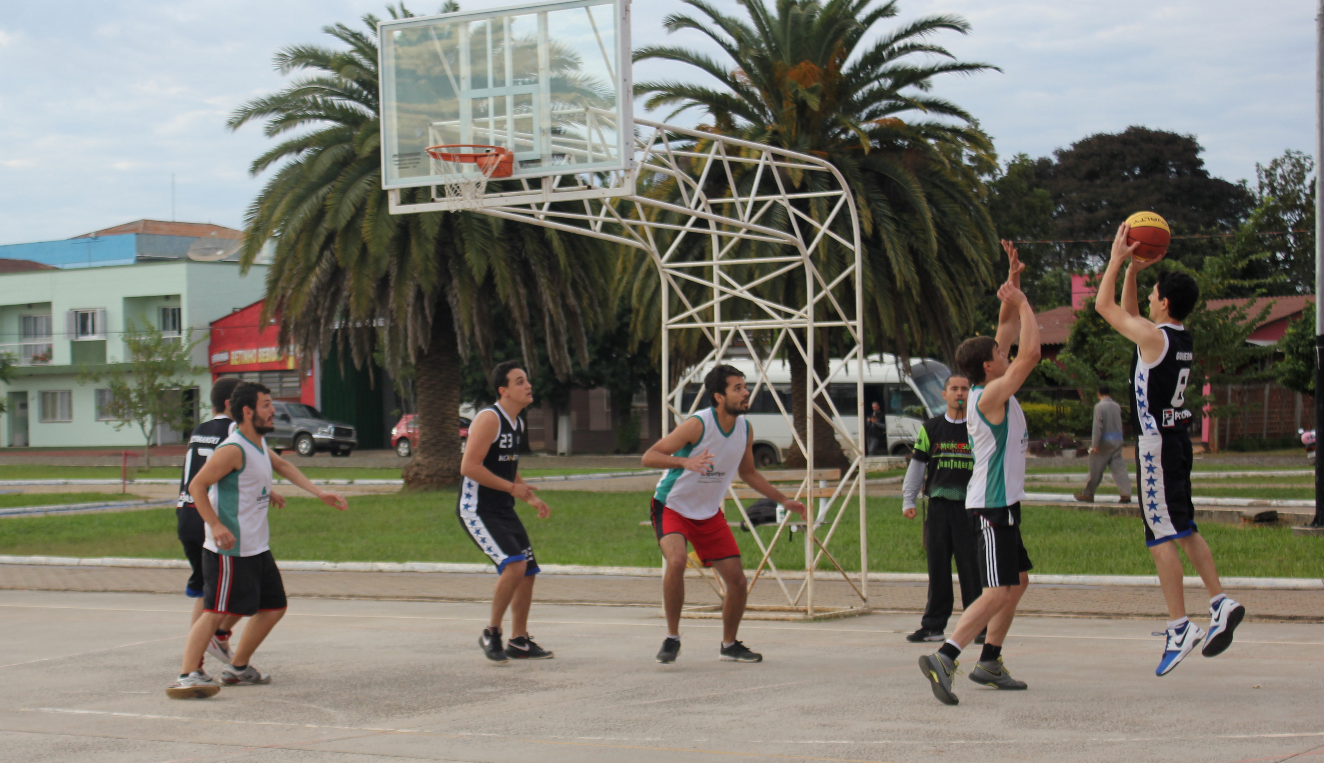 seis jogadores, três de cada time, jogam basquete em uma quadra de cimento; à esquerda , um jogador arremessa a bola rumo à cesta, enquanto os demais marcam seus adversários e o rumo da jogada