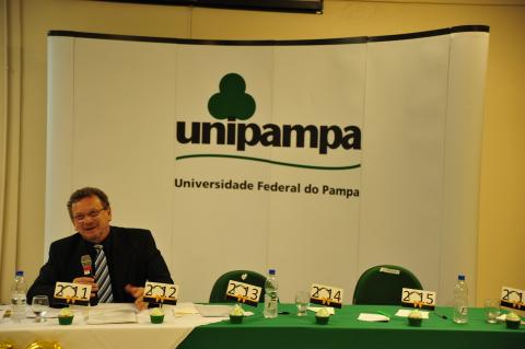 O deputado está de terno escuro, sentado, em frente ao painel com o nome da Unipampa, fala para a plateia sobre o papel da Unipampa no desenvolvimento da região.