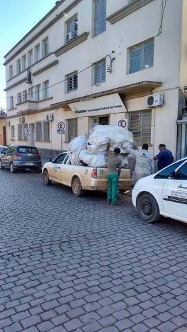 Na fotografia, três homens carregam a caminhonete da Prefeitura de Bagé com a carga de materiais que deverão ser reciclados, em frente ao prédio da Unipampa.