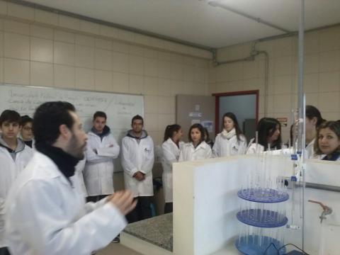 Grupo de pessoas jovens reúne-se, todos de jaleco branco e posicionados em pé, em torno de bancadas com equipamentos do laboratório de química.