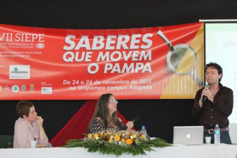 duas mulheres estão sentadas à frente da mesa de honra, e olham para um homem que fala ao microfone. Ao fundo, um banner com a marca e o nome do evento